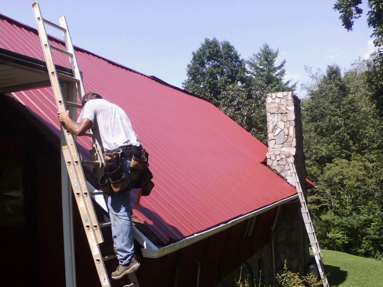 Roofer on a ladder beside a red metal roof and stone chimney — Soffit & Fascia Repair by Roofing Brothers in Sellersville